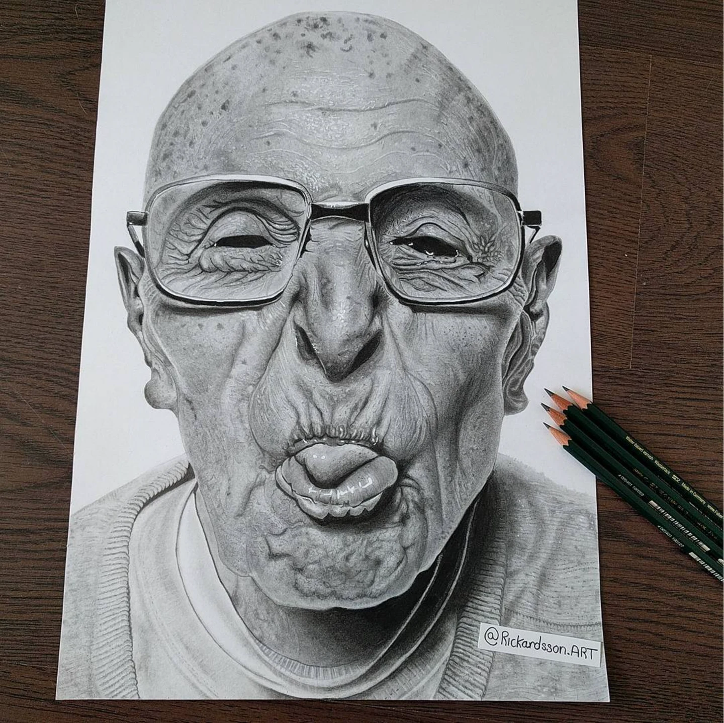 Drawing of an elderly man wearing glasses and sticking out his tongue, on paper beside pencils.